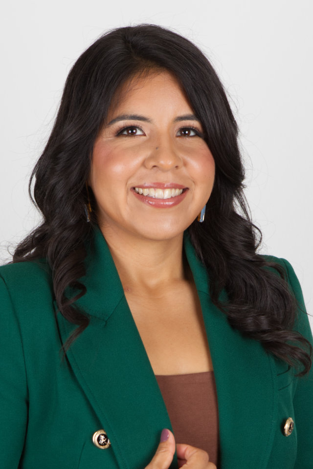 Woman with long dark hair wearing a green blazer and brown top, smiling at the camera against a plain white background.