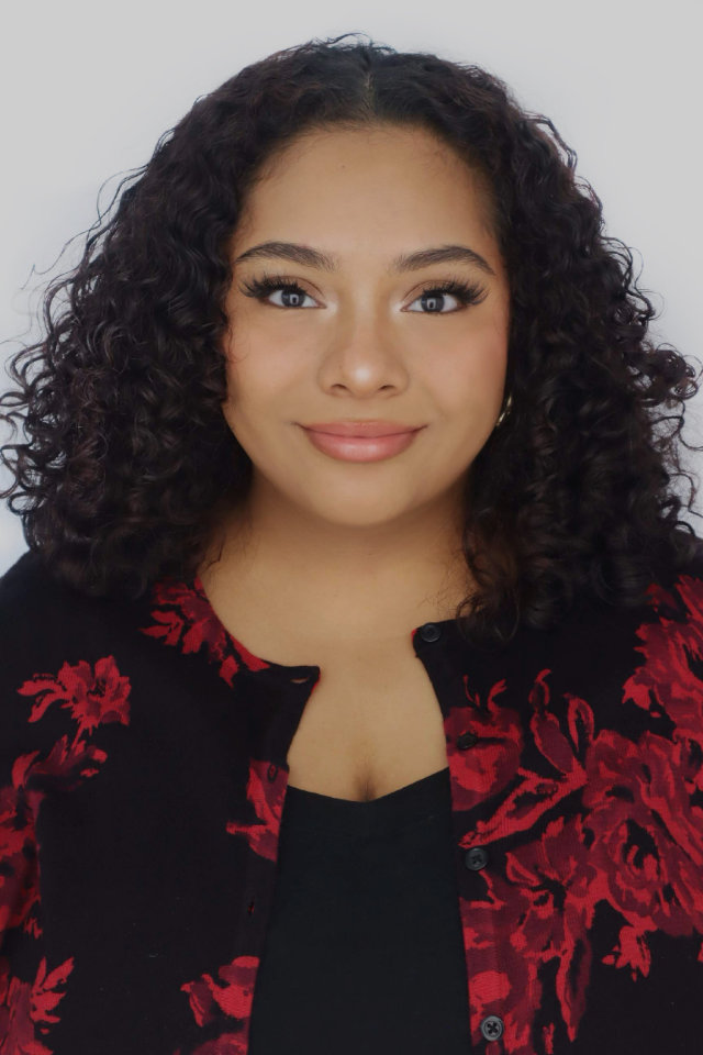 A woman with curly dark hair and natural makeup, wearing a black top and a black cardigan with a red floral pattern, poses against a plain light background.