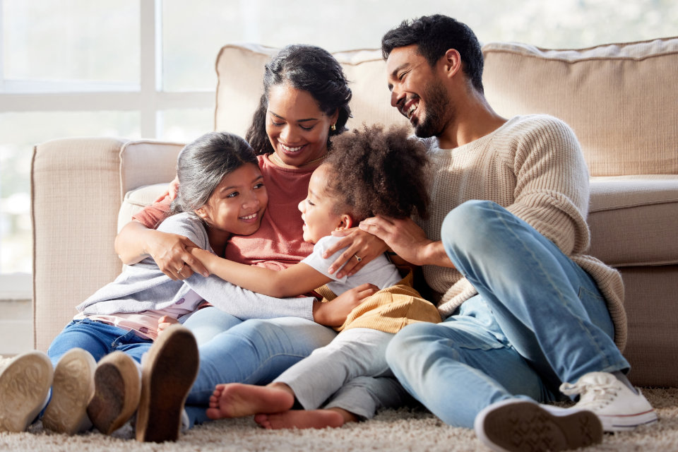 A family of four sits on the floor in front of a couch, smiling and hugging each other in a brightly lit room.