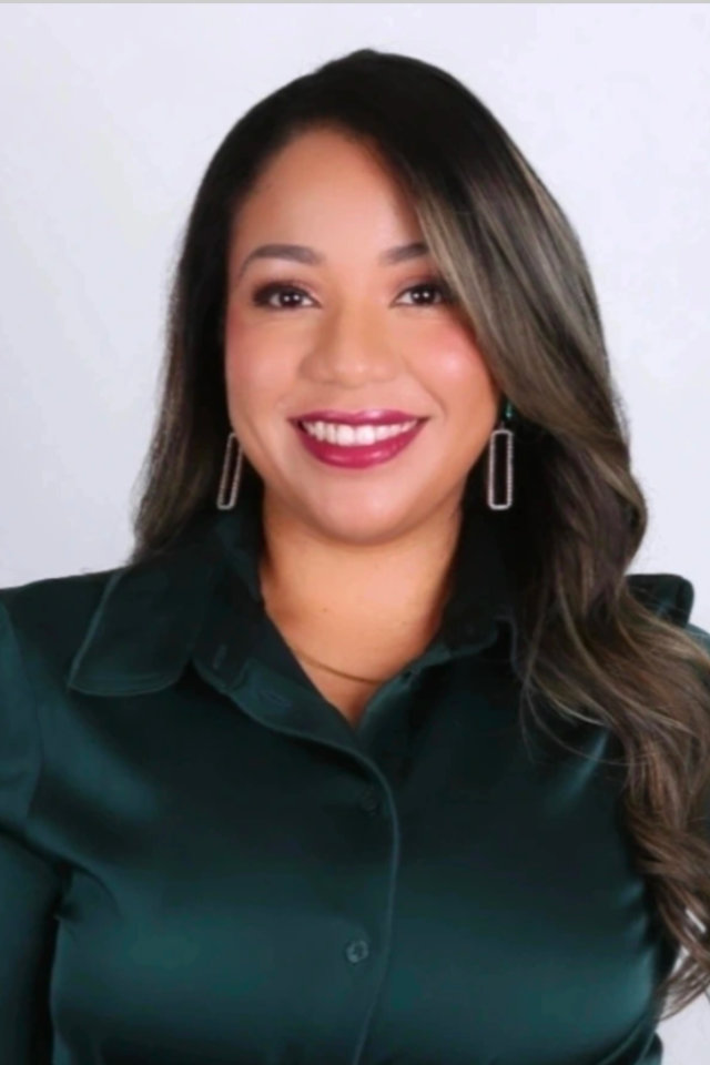 A woman with long, wavy brown hair, wearing a dark green blouse and geometric earrings, smiles in front of a plain light background.