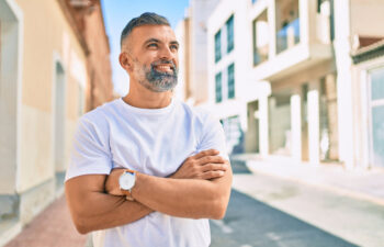 Man with gray hair and beard wearing a white t-shirt stands outside on a sunny day with arms crossed, smiling, in an urban street setting.