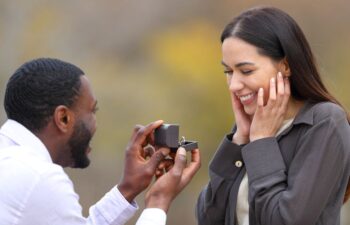 An Afro-American holding an open cox with an engagement man and proposing to a happy, surprised Hispanic woman
