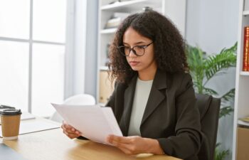 A woman with curly hair and glasses sits at a desk, reading documents. There is a coffee cup, bookshelves, and a plant in the background.