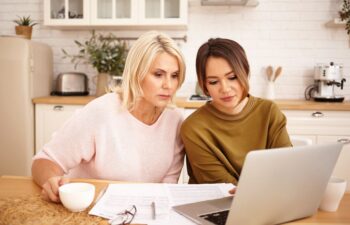 Two women sit at a kitchen table looking at a laptop, with papers and a cup in front of them.